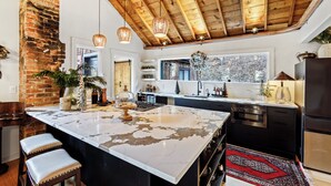 A modern kitchen with a large island, wooden ceiling beams, hanging pendant lights, and a view of the outdoors through a window. - Cozy river front 3 bedroom Colorado cabin (Colorado Springs)