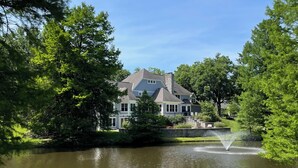 Une grande maison avec un étang et une fontaine à l’avant, entourée d’arbres.