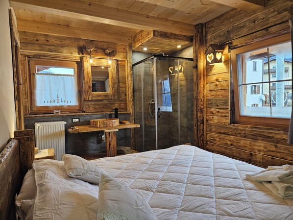 A wooden cabin-style bedroom with a bed, a vanity with a mirror, a shower, and a window with a view of a building. - Chalet between Cortina d'Ampezzo and the Marmolada, Tabià Falesia  (Laste di Sopra)