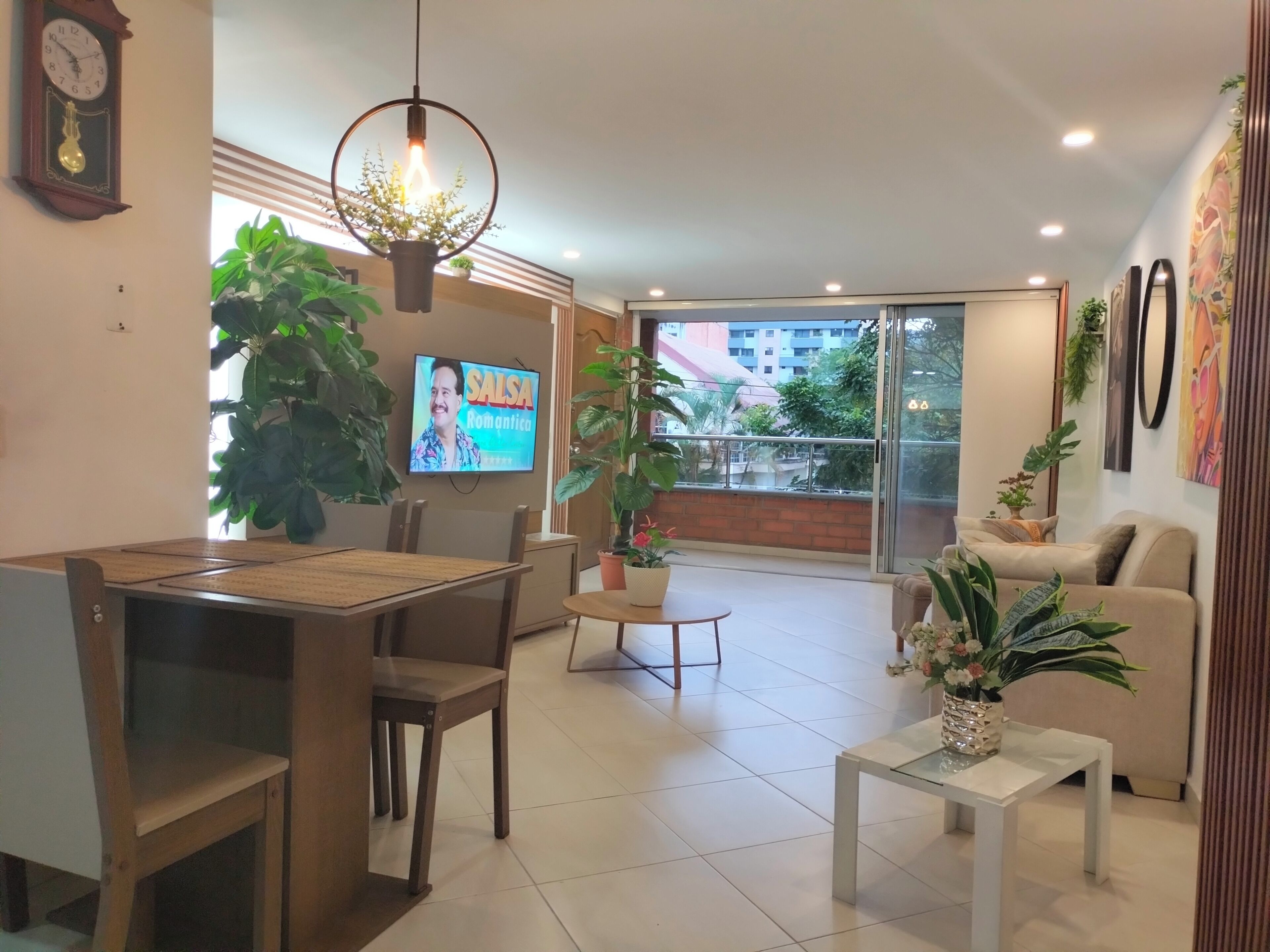 A modern living room with a dining area, large windows, and a TV displaying 'Salsa Romantica.'
