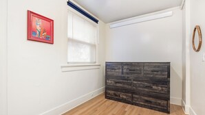 Ein Zimmer mit Holzboden, einem dunklen Holzschrank, einem Fenster mit Jalousien und einem gerahmten Bild an der Wand.