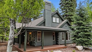 Une cabane en rondins avec un porche, une cheminée et un grand arbre à feuilles persistantes en arrière-plan.
