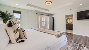 A bedroom with a large bed, a television mounted on the wall, and a door leading to a bathroom. - The Palm Hideaway Oasis with Pool + Hot Tub + Fire pit & Outdoor kitchen! (Spicewood)