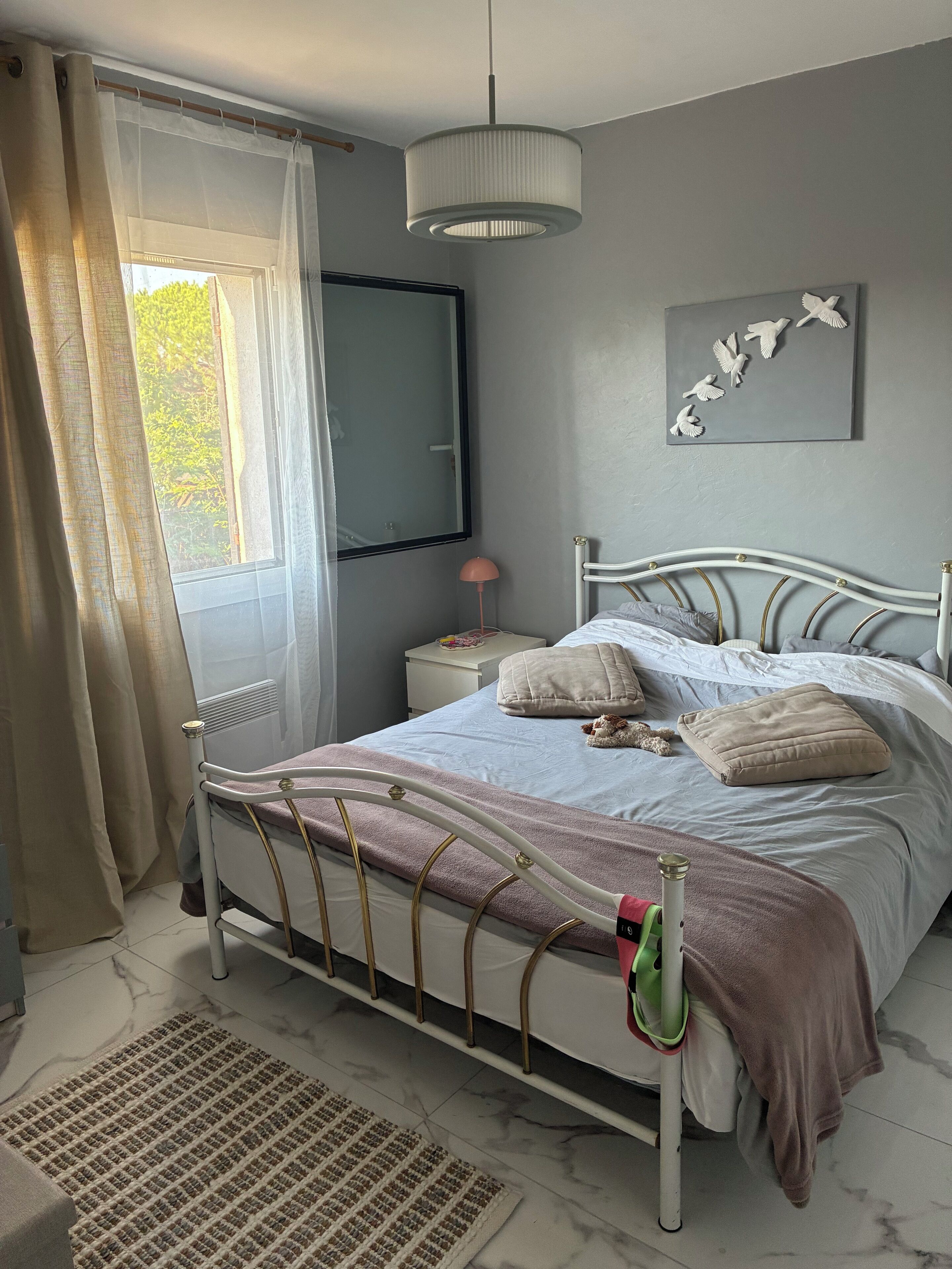 Ein Schlafzimmer mit Bett, Nachttisch, Lampe, Heizkörper, einem Fenster mit Vorhängen und einem Wandbild.