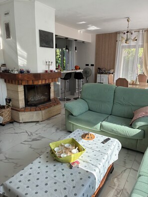 A living room with a fireplace, a green sofa, a dining table with a polka dot tablecloth, and a kitchen area in the background. - Charming House by the Sea and Near the Janas Forest – Quiet & Cozy 5-Bedroom Home - (La Seyne-sur-Mer)