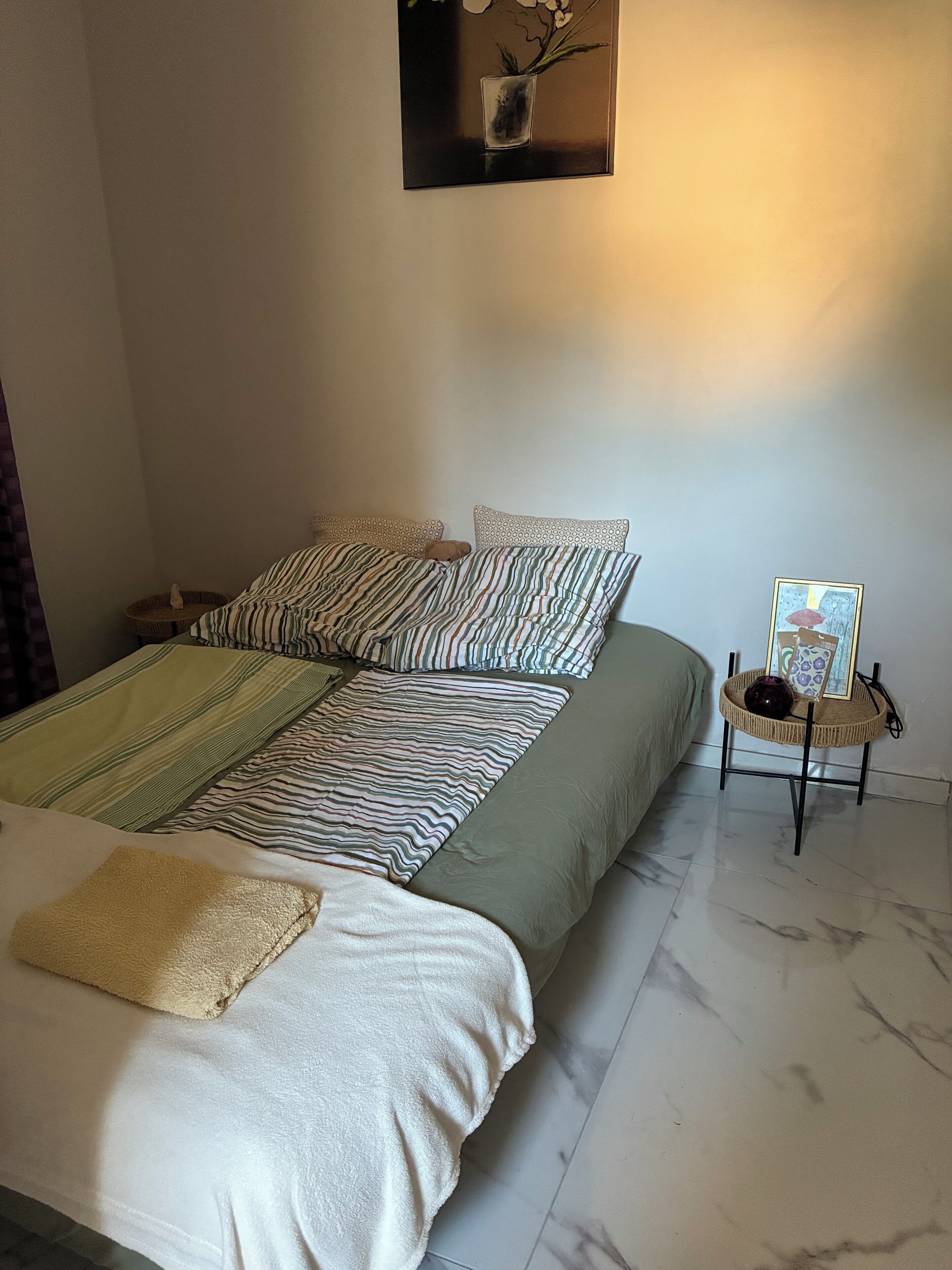 A neatly made bed with striped bedding, a small bedside table with a vase and picture frame, and a wall-mounted picture above the bed.