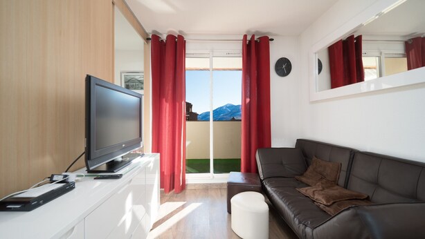 Ein modernes Wohnzimmer mit Flachbildfernseher, braunem Ledersofa, roten Vorhängen und Bergblick durch eine Glastür.