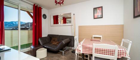 Ein modernes Wohnzimmer mit einer Couch, einem Esstisch mit Stühlen, einer rot-weißen karierten Tischdecke, Holzboden, einer roten Chandelierleuchtre und einer Wanduhr sowie einem gerahmten Bild an der Wand.