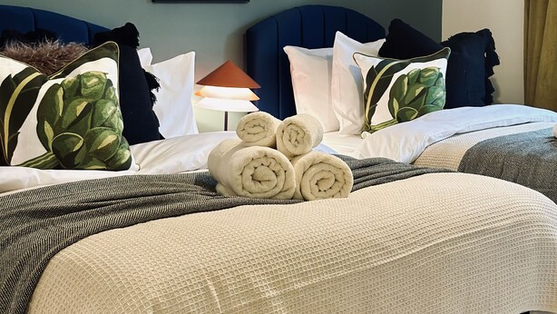 A hotel room with two beds, each with a dark headboard, white bedding, and decorative pillows. There are rolled towels on the beds, and a lamp on the nightstand. - Spacious Home | Free Parking | WiFi | Sleeps 8! (Gravesend)