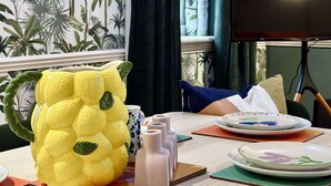 A dining area with a table set for two, featuring plates with floral designs, a yellow pineapple-shaped cushion, and a wall with a tropical wallpaper. - Spacious Home | Free Parking | WiFi | Sleeps 8! (Gravesend)