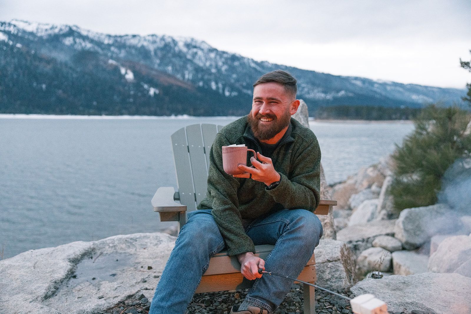 A person sitting on a wooden chair by a lake with mountains in the background.
