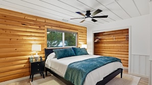 A bedroom with a wooden wall, a bed with a white and teal bedspread, a black nightstand, a white lamp, and a ceiling fan. - Beach Access + Hot Tub | Near Tamarack & McCall (Donnelly)