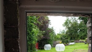 Blick durch eine Tür in eine grüne Wiese mit zwei großen, transparenten Kugeln, einer Steinmauer und einer Schaukel.