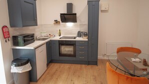 Eine kompakte K üche mit grauen Schränken, einer Mikrowelle, einem Spülbecken und einem kleinen Backofen. Ein Glastisch mit einem Stuhl und ein rotes Feuerlöschgerät sind an der Wand angebracht.