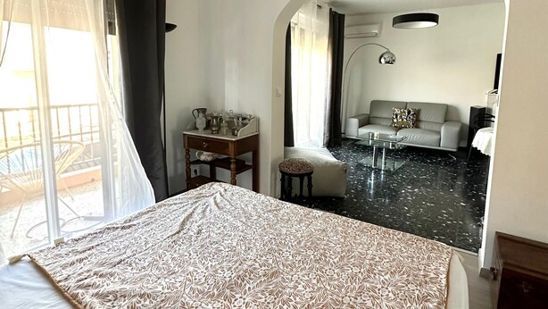 Une chambre d’hôtel avec un lit, une petite table avec une lampe, un canapé et une porte en arc donnant sur une autre pièce.