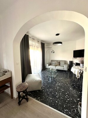 Ein Wohnzimmer mit einer Couch, einem Glastisch, einem Esstisch mit Stühlen, einem Fernseher und einer Deckenleuchte.