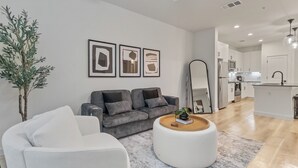 Een moderne woonkamer met een grijze bank, een witte fauteuil en een salontafel. Er hangt een grote spiegel aan de muur en staat een potplant. De kamer loopt over in een keuken met witte kasten en een centraal eiland.