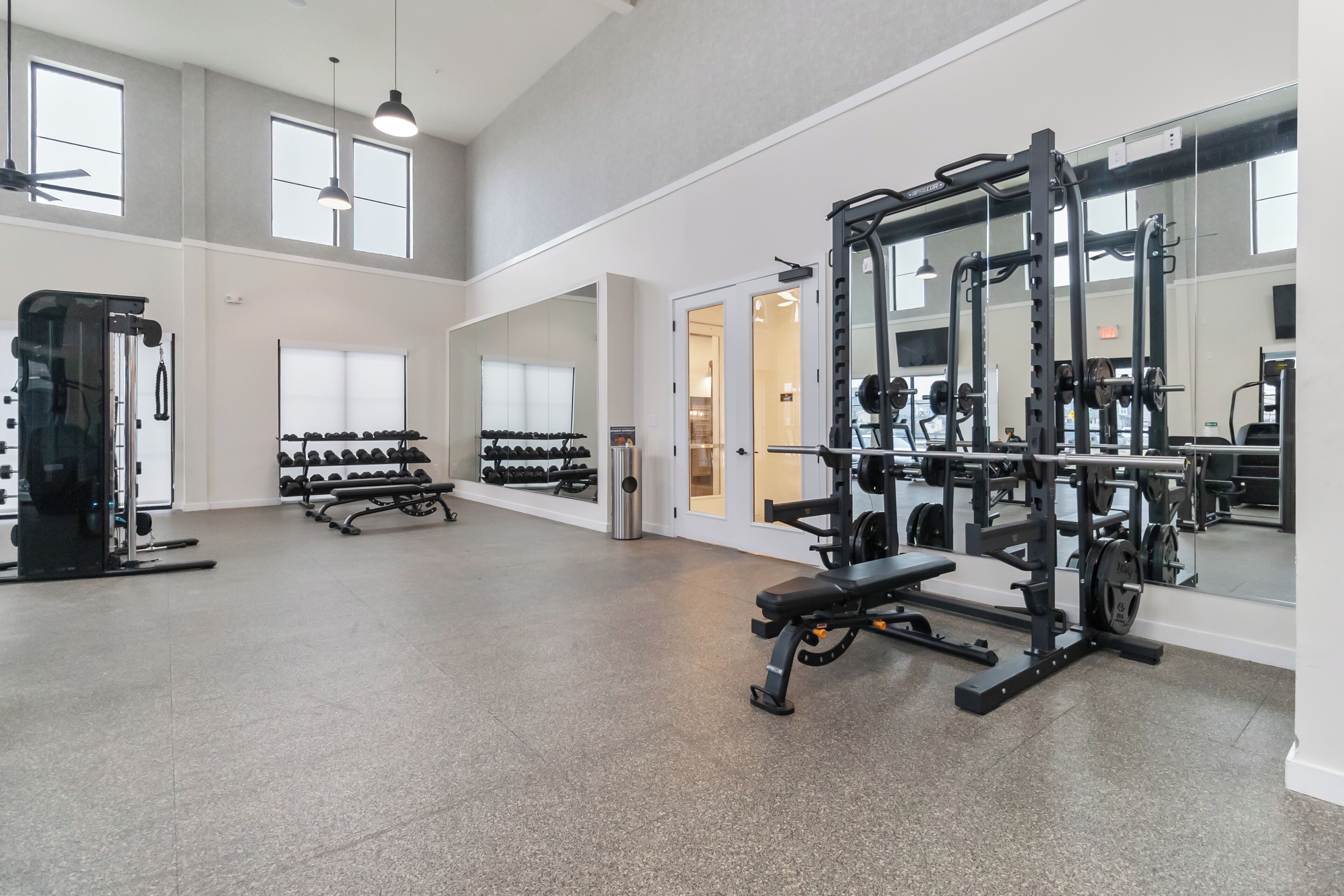 A spacious gym with various exercise equipment, large windows, and a high ceiling.