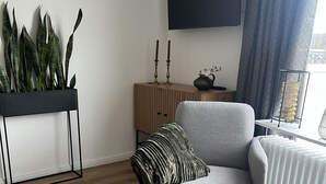 Un soggiorno moderno con una poltrona grigia, un mobiletto in legno, una lampada da terra nera, una pianta in vaso e un televisore a schermo piatto fissato alla parete.