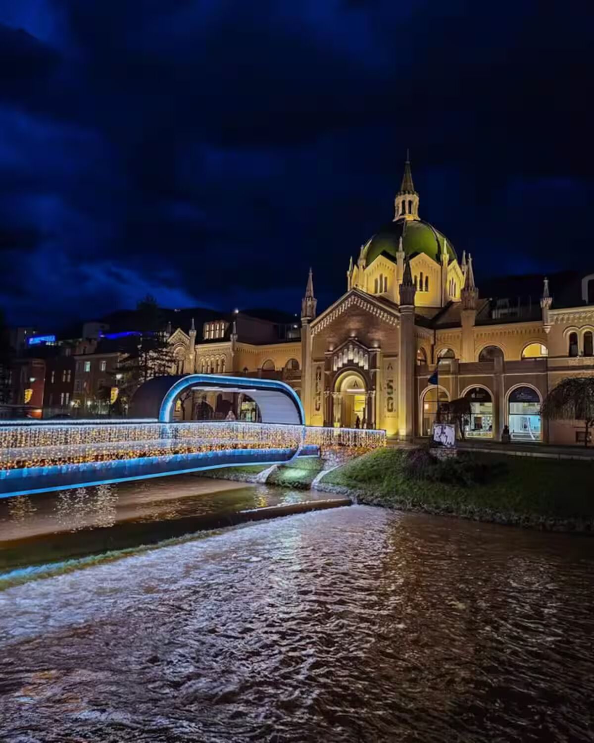A historic building with a large dome and arched windows, a bridge with blue lights, and a calm river in the foreground.