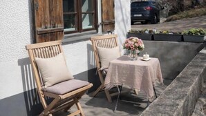 Ein Gebäude mit weißen Wänden und hölzernen Fensterläden, zwei Holzstühle mit Kissen, ein kleiner Tisch mit rosa Tischdecke und eine Blumenvase.