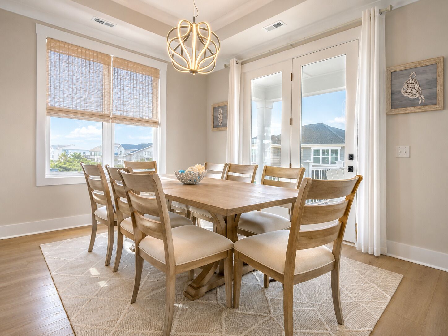 Comedor con una mesa y sillas de madera, una araña y vistas a casas a través de amplios ventanales.