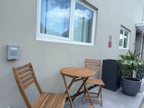 Ein kleiner Außenbereich mit zwei Holzstühlen und einem runden Tisch, einer Topfpflanze und einem an der Wand montierten Stromkasten.