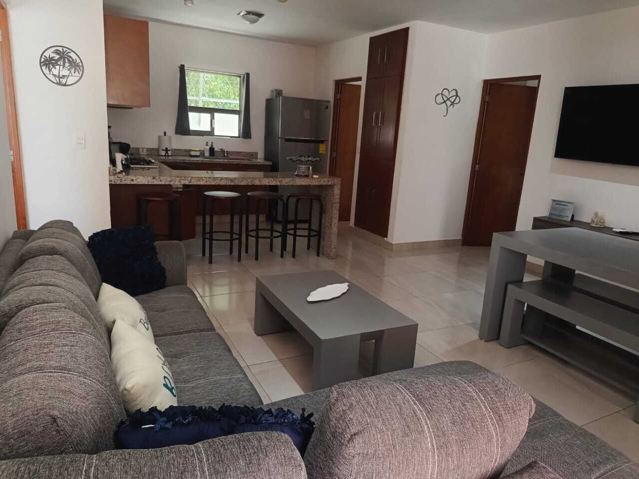 Una sala de estar con un sofá, una mesa de centro y un televisor. Hay una cocina con un refrigerador y un ventanal.