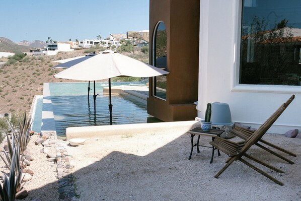 Un espace piscine avec une chaise et un parasol, entouré d’un paysage sablonneux et offrant une vue sur des bâtiments éloignés.