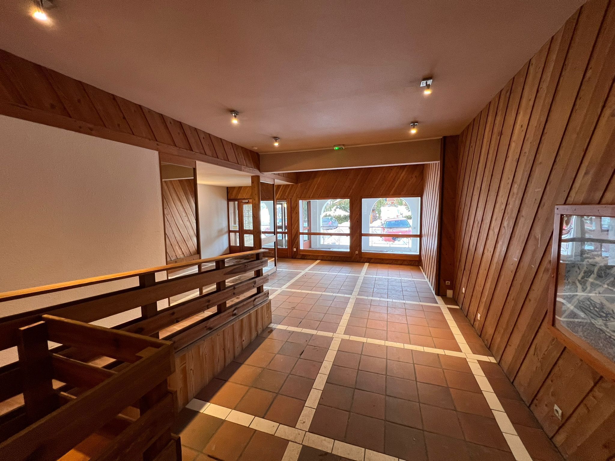 A spacious hallway with wooden paneling and a tiled floor, leading to large windows with a view of parked cars outside.
