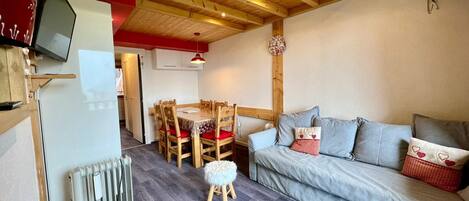 A cozy living room with a wooden ceiling, a grey sofa with patterned cushions, a wooden dining table with chairs, and a small stool.