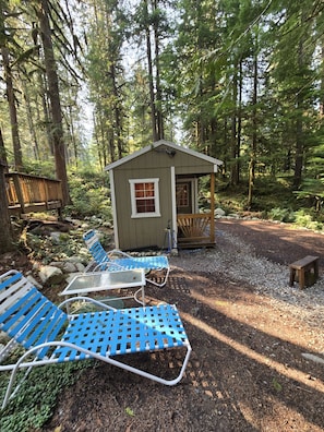 A cabin with a porch, surrounded by a forest. There are metal chairs and a table on the ground.