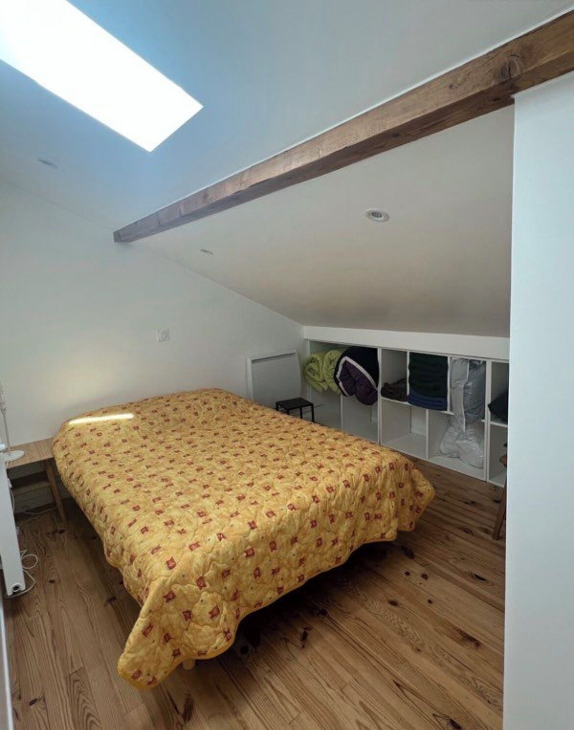 Une chambre à coucher avec un lit, un plancher en bois, des murs blancs, un puits de lumière et une armoire intégrée.