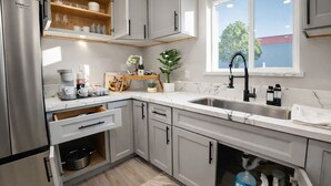 Een moderne keuken met grijze kastjes, een roestvrijstalen koelkast, een marmeren aanrechtblad, en een raam met uitzicht op bomen en een gebouw.