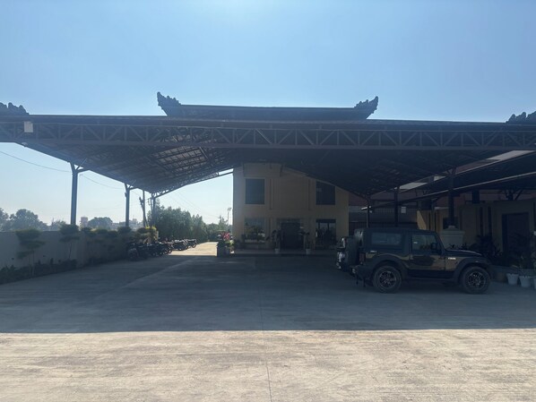 A parking area with a covered structure, a building with a flat roof, and a vehicle. - Ramaya Motel & Resorts (Ujjain)