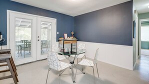 Een eetgedeelte met een glazen tafel en draadstoelen, een houten buffetkast en een deur naar een terras.