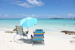 Two beach chairs and a blue umbrella on a sandy beach with clear turquoise water and a distant shoreline. - Large tropical group retreat | Beach just steps away | Spacious modern home (Nassau)