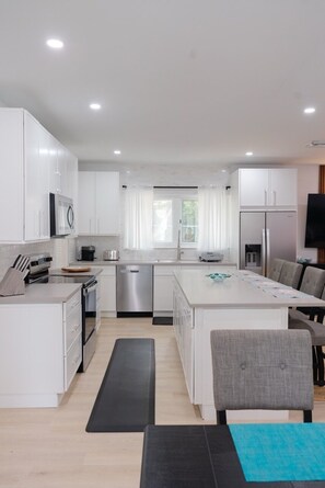 Une cuisine moderne avec des meubles blancs, un îlot central et un coin repas agrémenté d’une chaise grise.