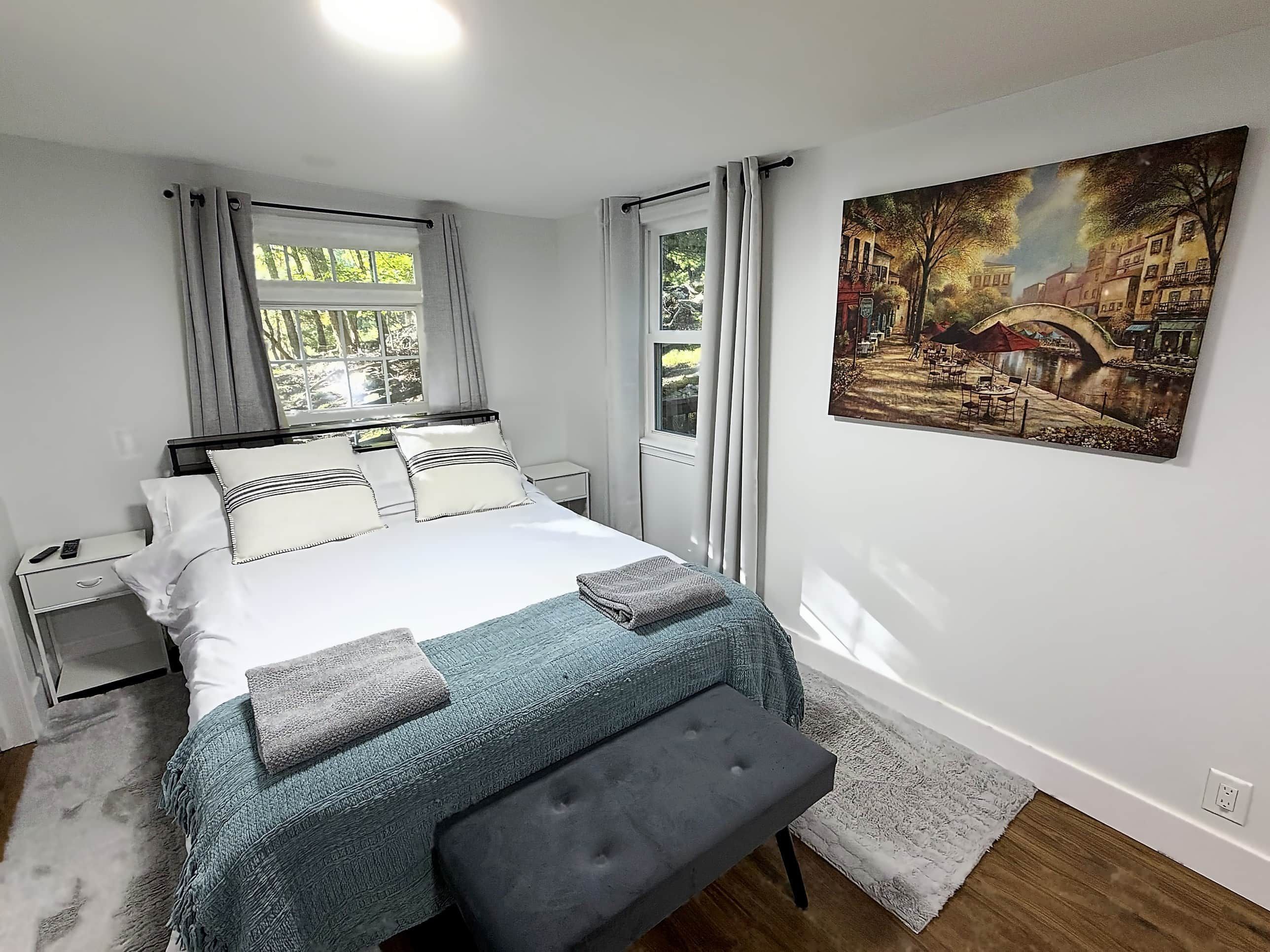 Ein Schlafzimmer mit einem Bett, Nachttischen, einer Bank, einem Gemälde und einem Fenster mit Vorhängen.
