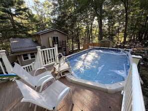 Une piscine avec une terrasse en bois, des chaises longues et une petite maison située dans un espace boisé.