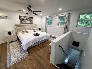 A bedroom with a bed, bedside tables, a ceiling fan, a fireplace, and a view of greenery outside. - Heated Pool, Fire Pit & Forest Views Poconos (Palmyra Township)