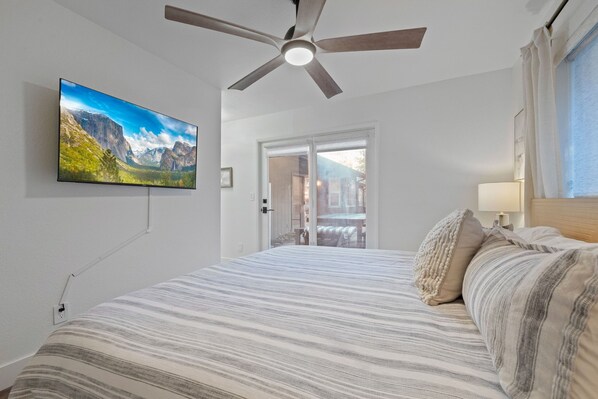 A bedroom with a bed, a ceiling fan, a TV mounted on the wall, and a window with curtains.