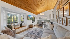A cozy living room with a wooden ceiling, large windows, a fireplace, and a dining area.