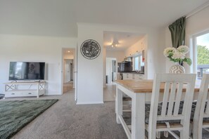 A modern living room with a TV, a dining area with a wooden table and chairs, a kitchen in the background, and a decorative wall piece. - Mango Point – Renovated Family Bach, Big Yard & Boat Parking a minute from Lake (Mangakino)