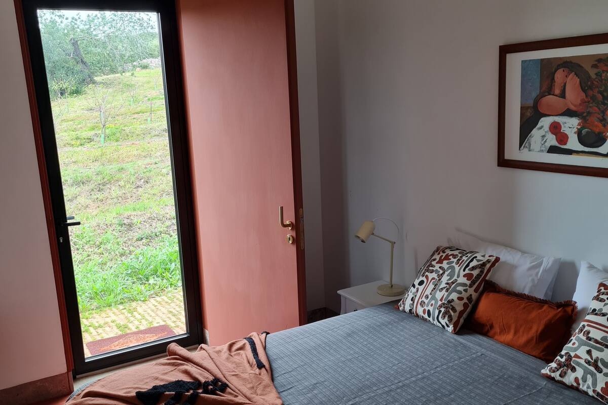 Een slaapkamer met een bed, een deur die uitkomt in een tuin, een nachtkastje met een lamp en een schilderij aan de muur.