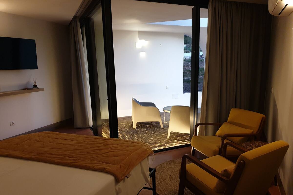 Een hotelkamer met een bed, een gele fauteuil, een klein tafeltje en uitzicht op het landschap.