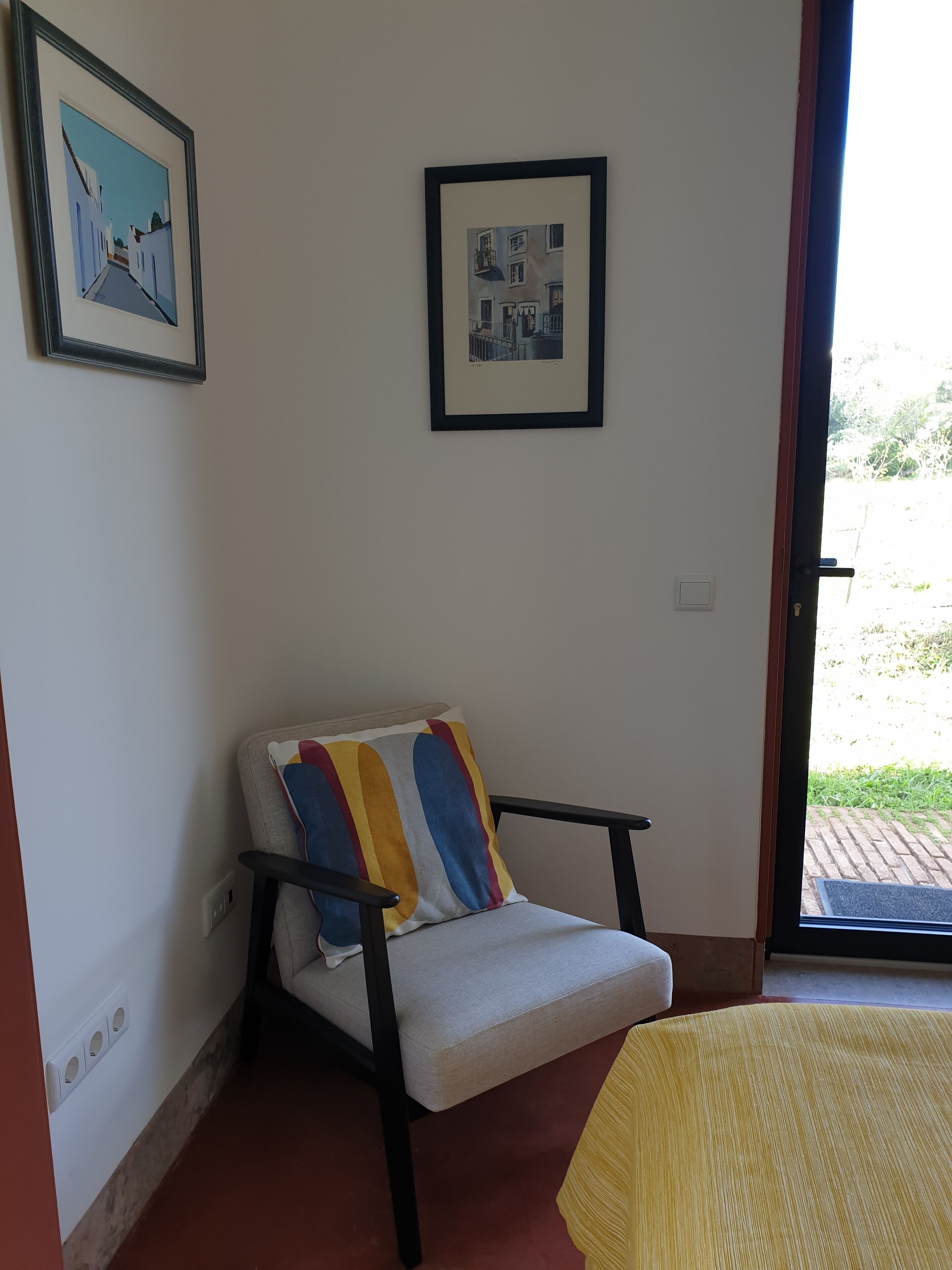 Een kamer met een stoel, een bed, een deur naar buiten en twee afbeeldingen in lijsten aan de muur.