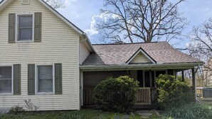 A two-story house with a covered porch, stone pathway, and a large tree in the background.