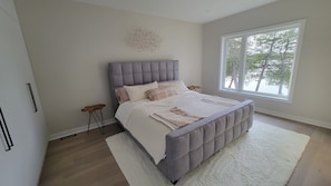 A bedroom with a tufted headboard, a large window, a white rug, and a bedside table with a lamp.