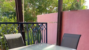 Ein Tisch mit Glasplatte und vier Stühlen steht auf einer gefliesten Terrasse mit Metallgeländer und einer rosafarbenen Wand im Hintergrund.